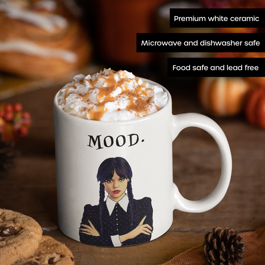 An 11 Oz white ceramic coffee mug featuring an illustration of Wednesday Addams with a deadpan expression. Below the image is the word “MOOD” in bold, black text. The design is printed on both sides, making it suitable for right- or left-handed users. A perfect gift for fans of dark humor and gothic style.