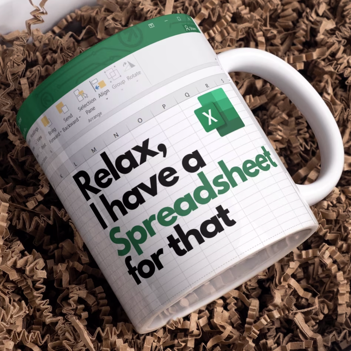 A white ceramic mug printed with a spreadsheet-themed design and the text 'Relax, I have a Spreadsheet for that,' shown resting on brown shredded paper.