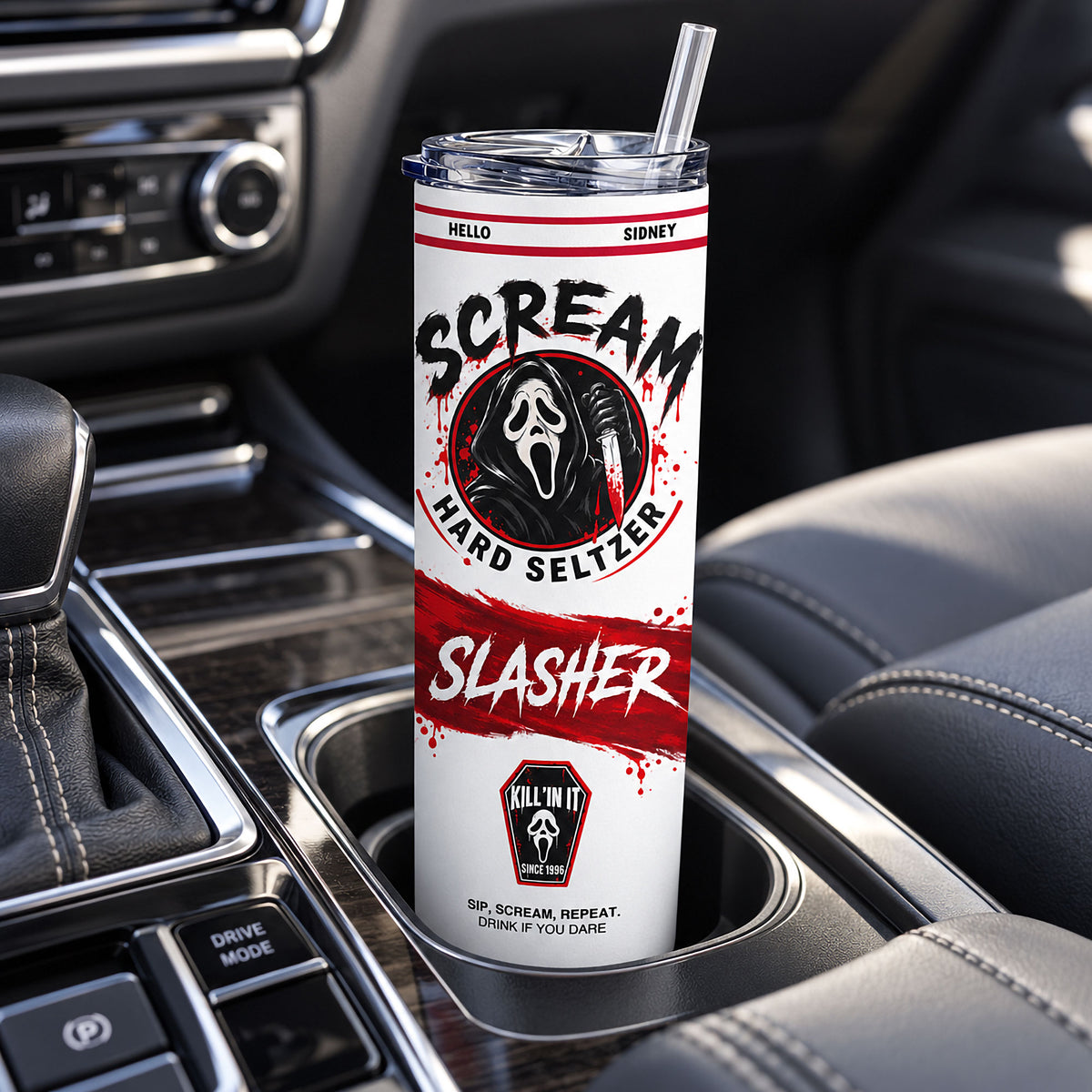 A white insulated tumbler with a clear lid and straw, printed with horror-themed 'Scream' graphics, placed in a car cup holder between seats.
