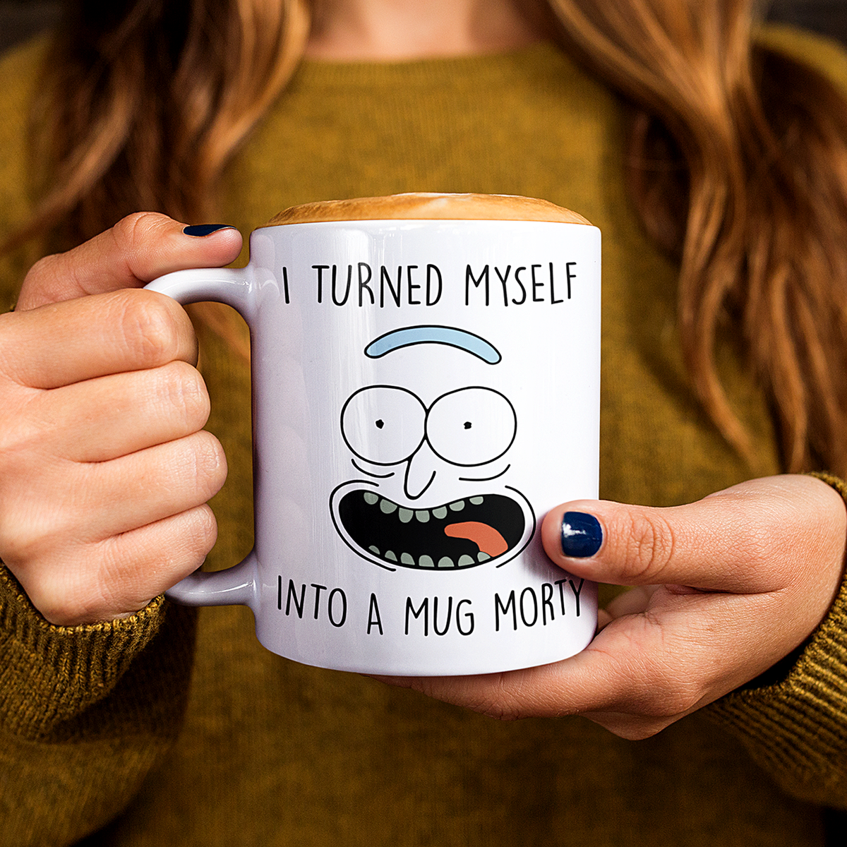 A person holding a white ceramic mug with a printed cartoon face and the text 'I TURNED MYSELF INTO A MUG MORTY'. The person is wearing a mustard green sweater, and the mug has a light wood-toned handle and rim.