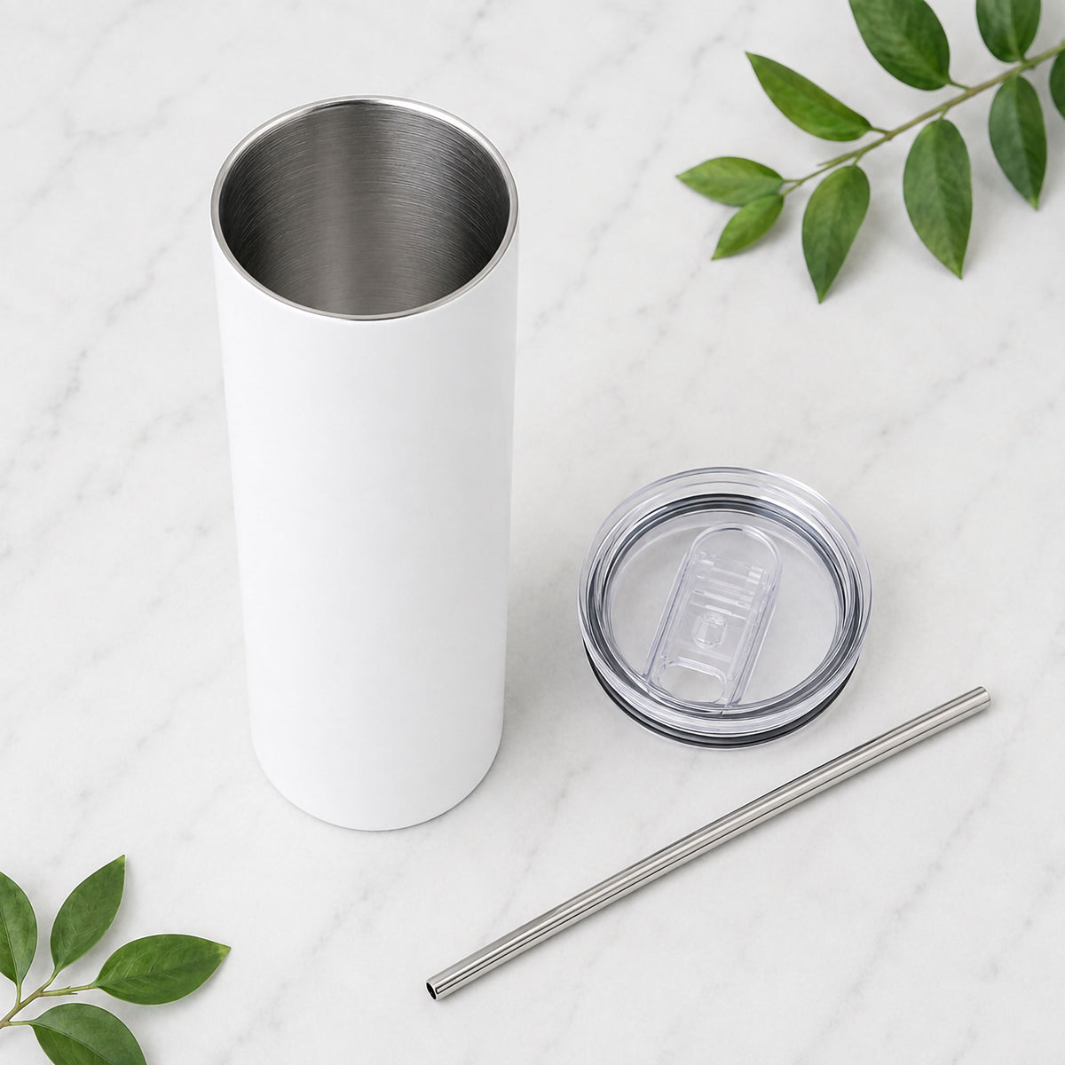 A tall white insulated tumbler with a stainless steel interior, shown with its clear lid and metal straw on a light marble surface with green leaves as props.